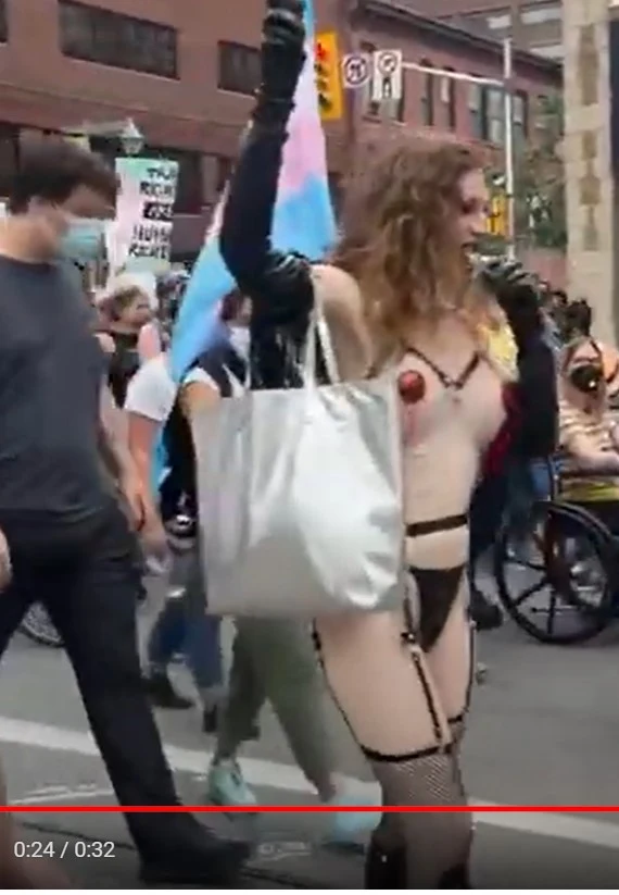 A video still (timestamp 0:24/0:32 shown on the playback bar) from an outdoor rally/march. A man with long curly hair walks through a crowd, topless except for a small red heart-shaped pasty, wearing black harness-style straps, a garter belt, and fishnet stockings, one arm raised holding a trans pride flag, the other holding a large silver tote bag. A man in a black shirt walks past in the foreground; other pedestrians, protest signs, and a person in a wheelchair are visible in the background.