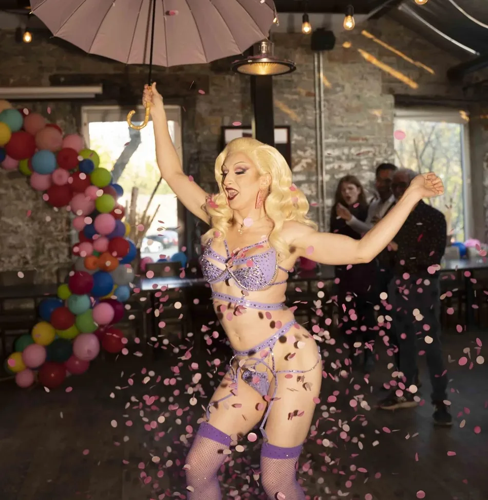 A photo of Tanya King, a burlesque performer, dancing indoors mid-performance, holding an open umbrella overhead with one hand and the other arm flung out, mouth open as if singing or shouting. He has long curled blonde hair, and wears a purple/lavender rhinestone-embellished bra-and-briefs set with matching thigh-high stockings. Pink and white confetti falls around him. Behind him, a large balloon garland (red, orange, yellow, green, blue, pink) frames the left side of the shot, string lights hang from the ceiling, and two onlookers stand near tables in the dim background, one appearing to clap.