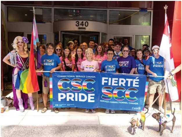 A group photo taken outside a building numbered "340," where roughly 20 people stand together for a Pride event photo. On the left, a drag performer (Tanya King, a man) in a purple outfit holds a rainbow flag; on the right, someone holds a Canadian flag. The group holds up a large banner reading "CORRECTIONAL SERVICE CANADA / CSC PRIDE" and "FIERTÉ SCC / SERVICE CORRECTIONNEL CANADA," with rainbow-colored lettering on "PRIDE" and "FIERTÉ." Two small dogs wearing rainbow bandanas sit in front of the group.