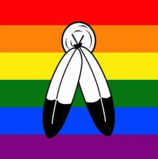 A flag featuring a rainbow background (red, orange, yellow, green, blue, purple stripes) with a black outline of two feathers crossed in the upper left corner over a circular sun/medicine-wheel-style emblem.