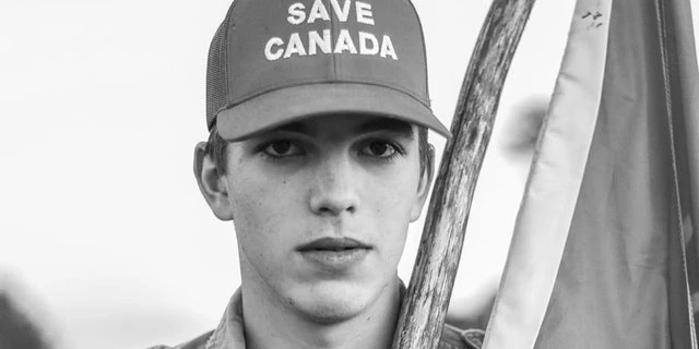 black-and-white close-up portrait of a young man looking directly at the camera, wearing a trucker cap reading "SAVE CANADA," holding a flagpole with a partially visible flag behind him.